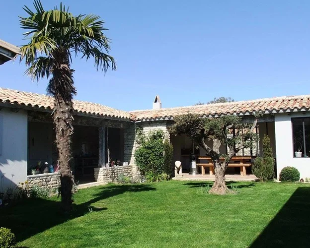 Cour ensoleillée avec pelouse verte, palmiers et bâtiments en pierre aux tuiles romaines à l'Ile de Ré en Charente-Maritime 17