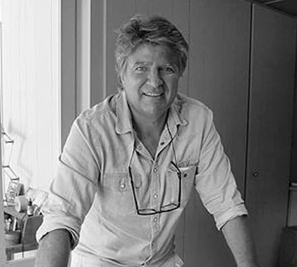 Homme souriant d'âge moyen, cheveux ébouriffés, lunettes suspendues à sa chemise claire.