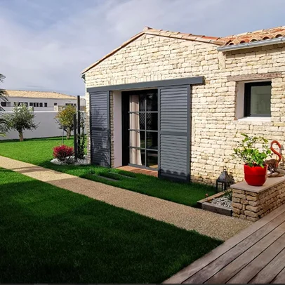Maison en pierre avec volets gris, jardin verdoyant, et chemin de gravier.