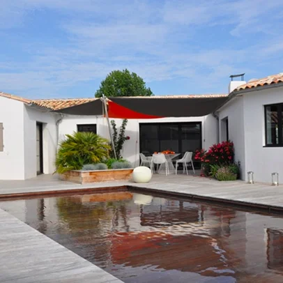 Piscine moderne reflétant un patio, avec des bâtiments blancs et un voile d'ombrage rouge.